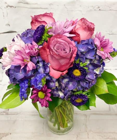 Let it rain...Purple Rain that is! Our most pleasing and popular purple floral design featuring sweetly scented lavender Roses, garden style Daisies and Hydrangea are designed together to create the perfect fullness in this mesmerizing purple tone-on-tone arrangement. We love the compact ball-shape tightness of the blooms together, making this an ideal floral design for any occasion, celebration, or the best of all, 'just because'.